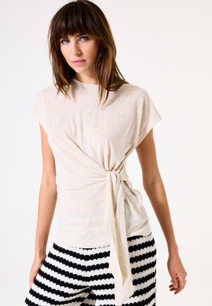 Haut beige clair à manches courtes avec un détail à nouer à la taille, fabriqué en tissu doux. Assorti à un short texturé noir et blanc à rayures.