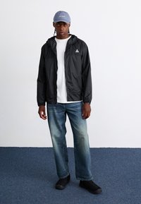 Veste noire légère avec capuche, t-shirt blanc, jeans bleus délavés et chaussures noires. Une casquette bleu clair avec un logo complète la tenue.