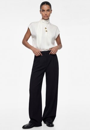 Femme aux cheveux foncés coiffés en chignon élégant, portant un chemisier blanc sans manches avec des boutons dorés, un pantalon large noir et des mocassins noirs, debout.