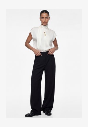 Femme aux cheveux foncés coiffés en chignon élégant, portant un chemisier blanc sans manches avec des boutons dorés, un pantalon large noir et des mocassins noirs, debout.