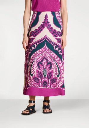 Femme portant une jupe longue à motifs roses avec des sandales noires et un haut magenta, debout devant un fond clair uni.