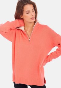 Pull en corail avec fermeture éclair, col montant, poignets côtelés et coupe décontractée. Le tissu semble doux avec des fentes sur le côté à l'ourlet pour plus de confort.