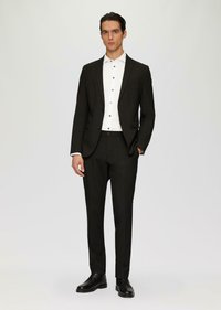 Costume noir avec une coupe ajustée, associé à une chemise blanche à boutons noirs. Texture uniforme et chaussures noires cirées.