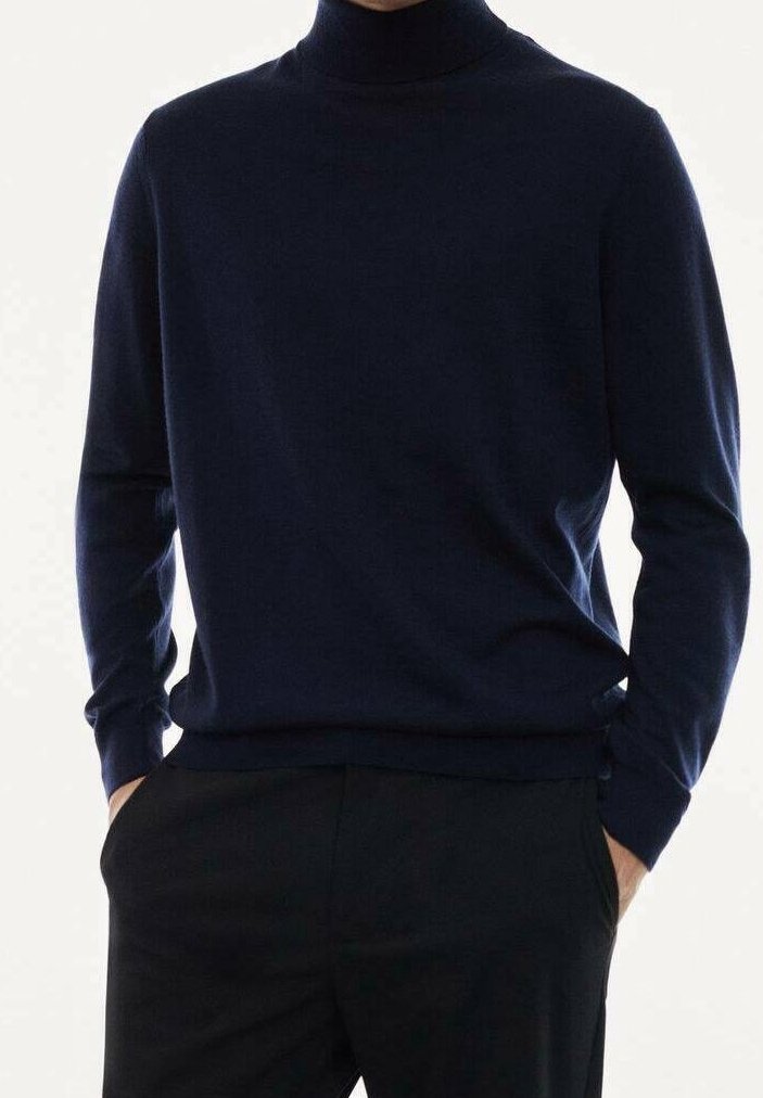 Ein Mann trägt einen dunkelblauen Rollkragenpullover, der leicht in schwarze Hosen gesteckt ist, mit den Händen in den Taschen vor einem hellen Hintergrund.