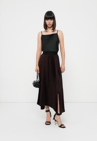 Camisola nera in satin abbinata a una lunga gonna plissettata bordeaux con uno spacco laterale, indossata con sandali neri con tacco alto e una borsa a tracolla di piccole dimensioni.