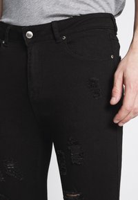 Jeans de mezclilla negra con parches desgastados y bordes deshilachados. Presenta botón y remaches plateados, diseño ceñido y ligera variación en la textura.