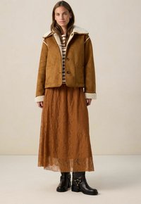 Bruine suedjas met schapenvacht kraag, gecombineerd met een roestkleurige kanten lange rok. Zwarte enkellaarsjes met gespen. Horizontaal gestreept topje zichtbaar.