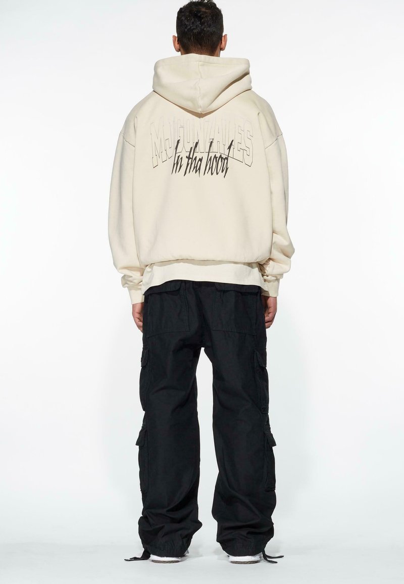 Beige oversized hoodie med "MOUNTAINS in tha hood" tryckt på baksidan, i kombination med svarta cargobyxor. Slät tyg, avslappnad passform.