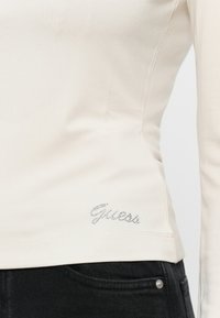 Kremowa koszula z długim rękawem, wykonana z miękkiego materiału, z srebrnym logo "Guess" w kryształkach na dolnym lewym boku. W zestawie z ciemnymi dżinsami.