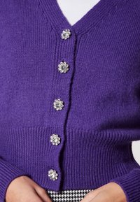 Lila Strickjacke aus geripptem Strickstoff, mit V-Ausschnitt und silbernen, blumenförmigen Knöpfen an der Vorderseite.