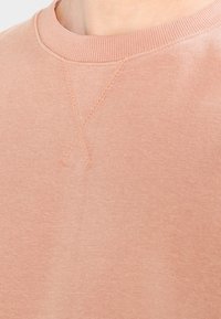 Sudadera color melocotón claro con cuello redondo acanalado, presenta un detalle sutil de punto cruz en la parte delantera y un tejido suave y texturizado.