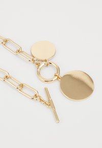 Zlatý řetízek s prodlouženými články, kulatými přívěsky a západkovým zapínáním. Hladký a lesklý povrch kovu s odrážejícími akcenty.