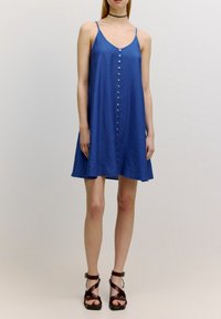 Robe bleue sans manches à coupe trapèze, en tissu texturé, avec fermeture à boutons sur le devant. Associée à des sandales marron à brides.