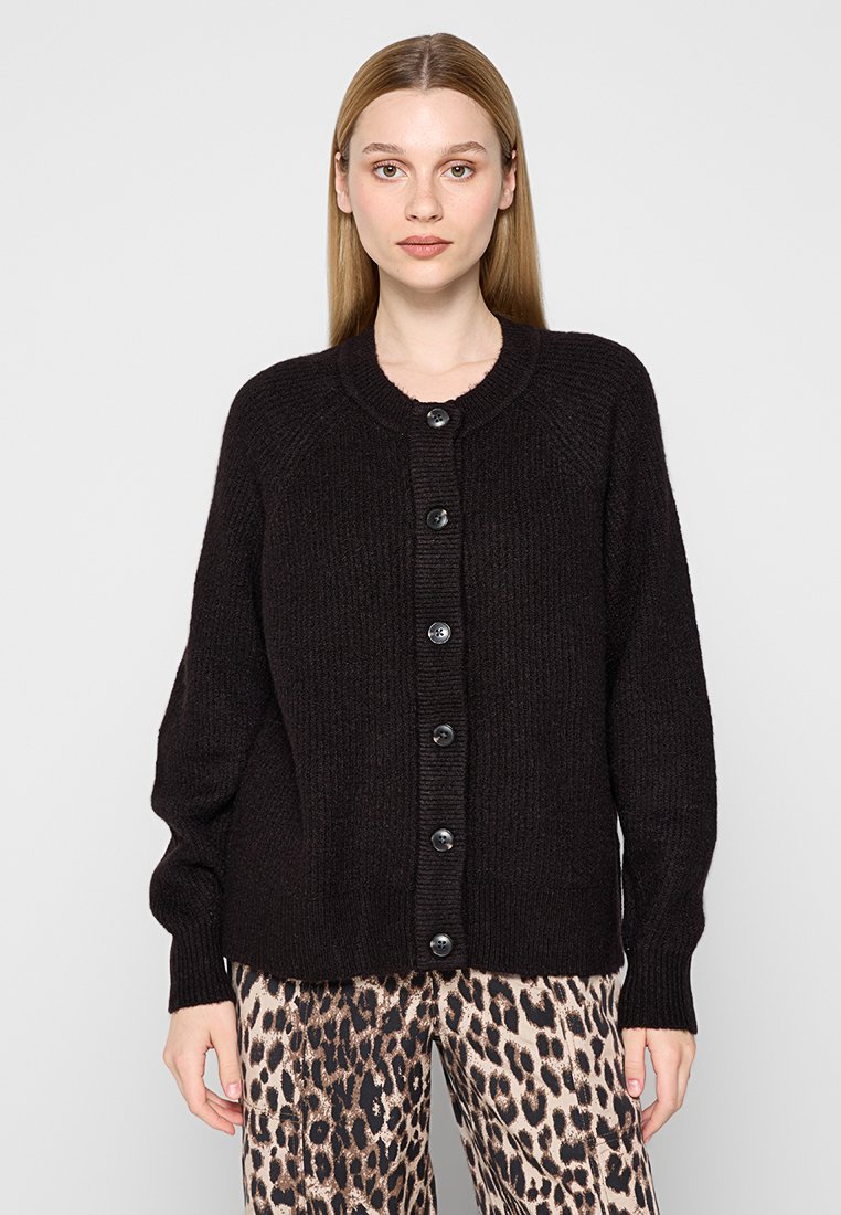 Schwarze Strickjacke mit Rundhalsausschnitt, ausgestattet mit einer knopfbaren Vorderseite und gerippten Bündchen. Getragen über einer Hose im Leopardenmuster.