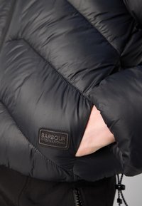Casaco puffer preto com um design acolchoado, apresentando um emblema da Barbour International, textura suave e um bolso com zipper.