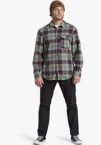 Geruite flanellen shirt in groene, paarse en blauwe tinten, met een knoopsluiting aan de voorkant en één borstzak; gecombineerd met zwarte broek en sneakers.