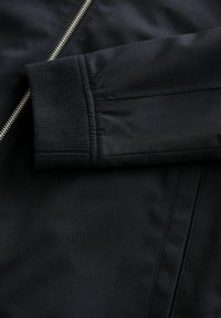 Chaqueta negra con una tela suave y ligeramente texturizada; cuenta con un cremallera y detalles de puños acanalados, enfatizando un diseño estructurado.