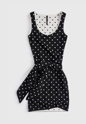 Robe noire sans manches à pois blancs, encolure dégagée, et ceinture en tissu nouée à la taille.