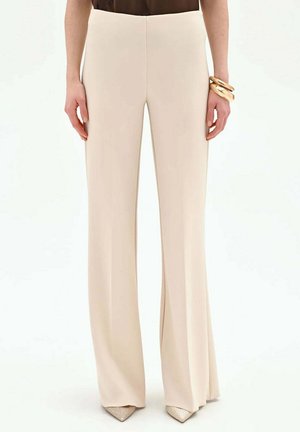 Persona che indossa pantaloni beige a vita alta e gamba larga, scarpe beige con punta, una maglietta nera e braccialetti grossi dorati al polso destro.