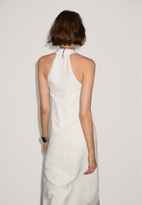 Robe blanche sans manches avec un col haut, présentant une silhouette ajustée et une texture subtile. Détail de fermeture éclair au dos visible.
