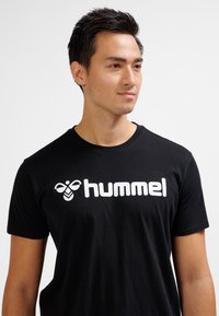 Ung man med kort mörkt hår som bär en svart t-shirt med en vit "hummel"-logotyp och en biikon, som tittar något åt höger mot en vit bakgrund.