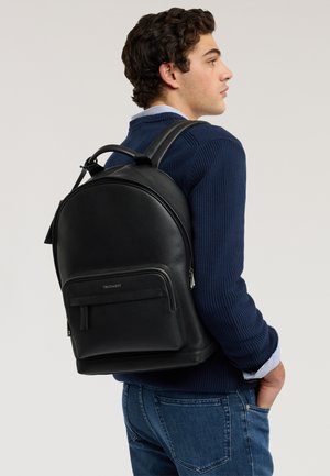 Junger Mann in blauem Pullover und Jeans, der einen schwarzen Leder-Rucksack mit Fronttasche und Tragegriff trägt, seitlich stehend.