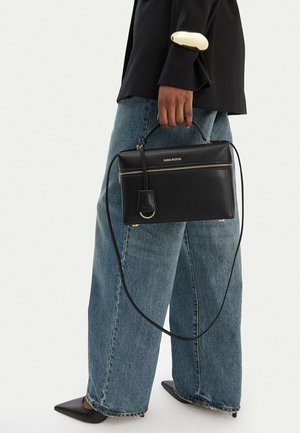 Persona che indossa jeans blu a gamba larga, scarpe nere con punta e blazer nero con dettagli dorati sui polsini, che tiene una borsa nera Gino Rossi.