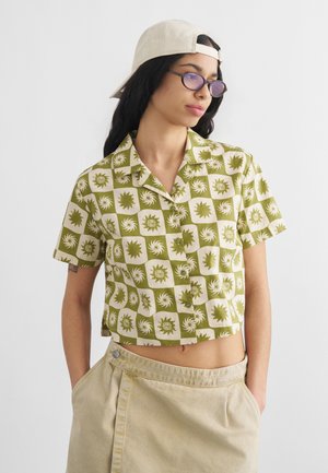 Młoda osoba ubrana w zielono-białą wzorzystą krótką koszulę, beżową spódnicę, beżową czapkę na odwrót, okulary z fioletowymi szkłami, z rękami w kieszeniach.