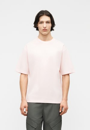 Giovane uomo con capelli castani di media lunghezza, indossa una T-shirt oversize rosa chiaro e pantaloni cargo grigio scuro su uno sfondo bianco uniforme.