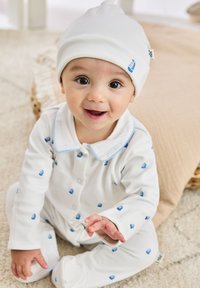 Witte katoenen rompertje met een blauw zeilbootmotief, drukknoopbevestigingen en blauwe bies op de kraag. Witte muts met een klein zeilbootaccent.