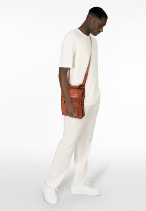 Braune Leder-Umhängetasche mit strukturiertem Oberflächendesign, mehreren Reißverschlussfächern und verstellbarem Tragegurt, gehalten von einer Person in einem hellen Outfit.