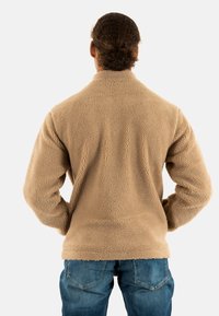Uomo con capelli ricci legati indietro, che indossa una giacca in fleece beige e jeans blu, visto di spalle su uno sfondo semplice.