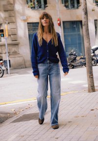 Blå cardigan med V-hals og knapper, kombineret med lyseblå vidde-bukser og brune sko, gående på en fortov.