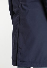 Marinblå vattentät jacka med rak passform, försedd med en central dragkedja, slät tygtextur och en diskret matt finish.