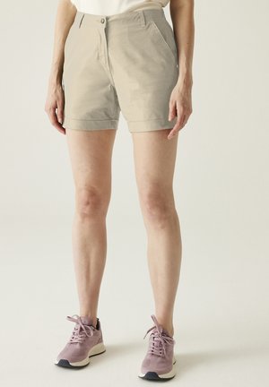 Persoon die beige korte broek en roze sportschoenen draagt, staat tegen een effen lichte achtergrond.
