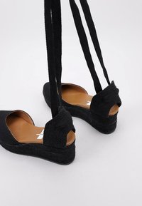Zapatos de esparto negros con cuña, con parte superior de tela texturizada, lados abiertos y correas entrelazadas. Plantilla marrón y suela texturizada negra.