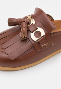 Chaussure marron en cuir à enfiler avec un design à pampilles à l'avant, un accent circulaire doré et une semelle texturée pour une meilleure adhérence.