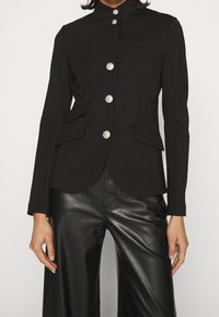 Chaqueta negra a medida con cuello alto, botones de plata y dos bolsillos laterales. Lleva puesta sobre pantalones negros brillantes, que presentan una silueta ajustada.