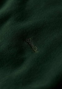 Tejido verde oscuro con una textura suave, que presenta un logo bordado "Silver 21" en hilo dorado sutil, ubicado en la parte inferior derecha.