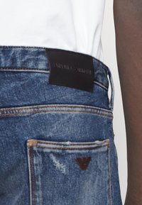 Džínové kalhoty v modrém odstínu s hnědou koženou etiketou s nápisem "EMPORIO ARMANI." Obsahují dvojité zadní kapsy a prošití.