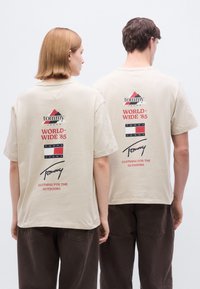 Μπεζ βαμβακερές μπλούζες διαθέτουν γραφικό στο πίσω μέρος με τις λέξεις "tommy JEANS," "WORLD-WIDE '85" και μια υπογραφή σε κόκκινες και μπλε αποχρώσεις. Κοντά μανίκια, άνετη εφαρμογή.