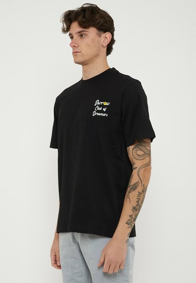 Young man with curly hair and sleeve tattoo wears black T-shirt with "Barrow Club of Dreamers" text and light gray pants.