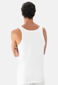 Mand med krøllet mørkt hår iført en enkel hvid tanktop, stående med armene over kors, vendt væk mod en lys ensfarvet baggrund.
