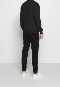 Svart sweatshirt med ribbade ärmslut och fåll, tillsammans med svarta mönstrade joggers. Vita sneakers kompletterar outfiten. Bilden visar bakifrån.