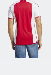 Mężczyzna w czerwono-białej sportowej koszulce z krótkim rękawem i niebieskich dżinsach, stojący tyłem z rękami luźno opuszczonymi wzdłuż ciała.