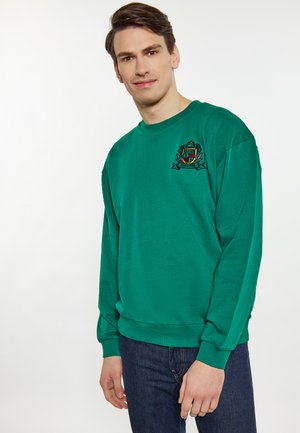 Grøn sweatshirt med lange ærmer, ribbede manchetter, rund halsudskæring og broderet crest-design på brystet. Bæres med mørke jeans.
