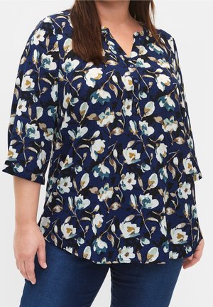 Femme portant un chemisier bleu marine à imprimé floral blanc et beige, manches trois-quarts, et un jean bleu sur un fond uni.