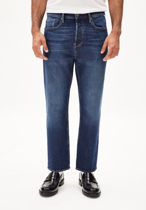 Dunkelblaue Jeans aus Denim mit geradem Schnitt, ausgestattet mit Vordertaschen, sichtbaren Nähten und kombiniert mit schwarzen Lederloafers.