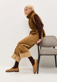 Bruine mouwloze trui, bruin longsleeve shirt, tan corduroy broek met brede pijpen en opgerolde mouwen, en bruine loafers. Stoel naast de figuur.