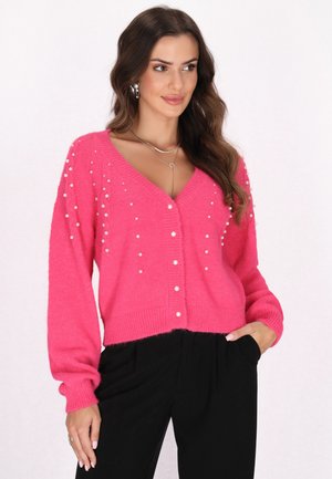 Cardigan roz dintr-un material moale, cu decolteu în V, accente de perlă și mâneci lungi. Stilizat cu pantaloni negri pentru contrast.